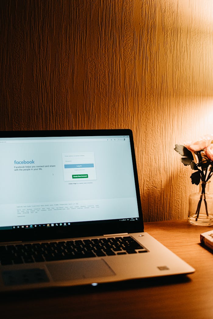 Warm-lit workspace featuring a laptop with Facebook on screen and a vase of flowers.